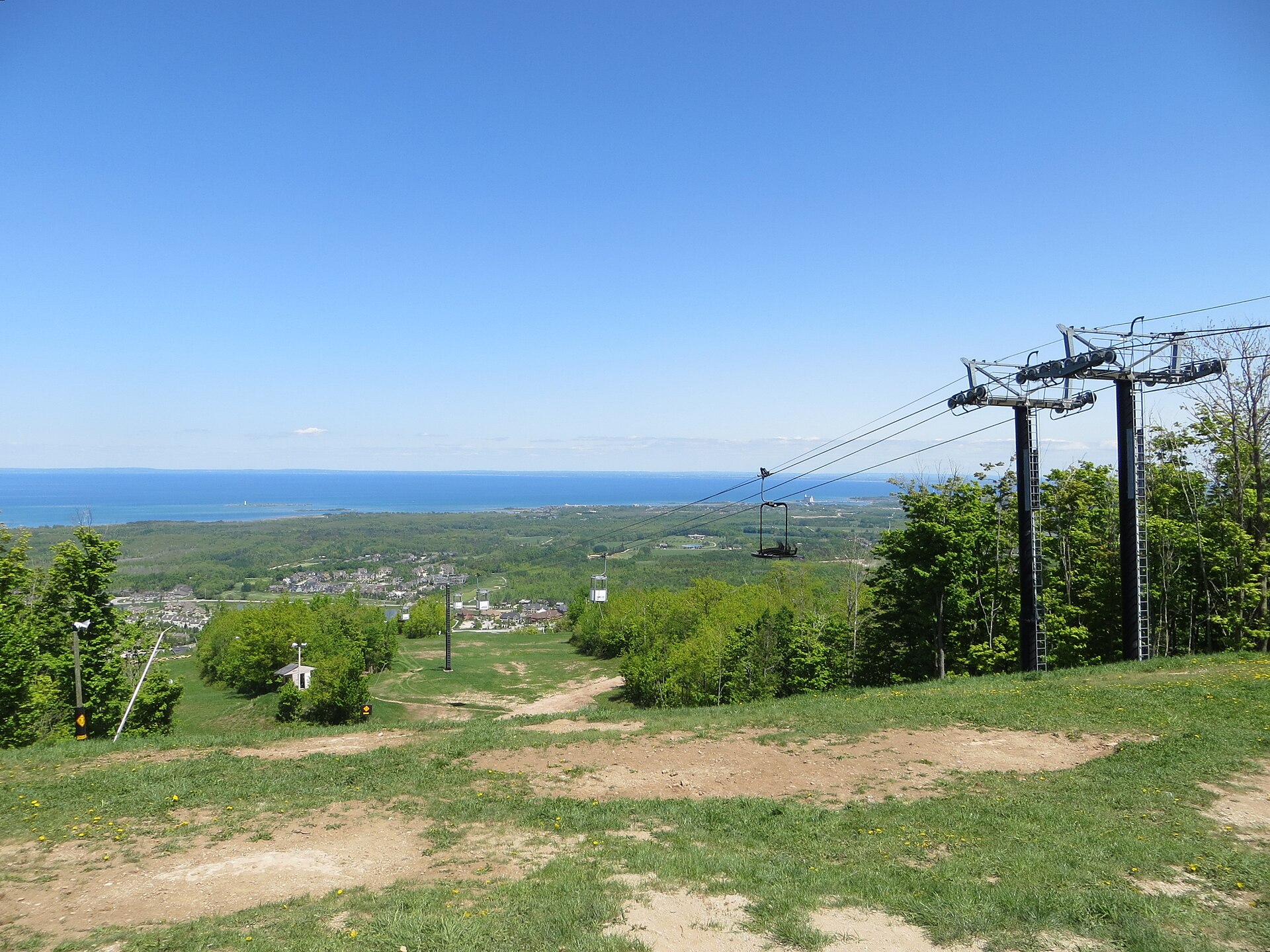 View from the top of Blue Mountain in Ontario down a grassy ski run and chairlift towards a village, green countryside and the blue water of Georgian Bay