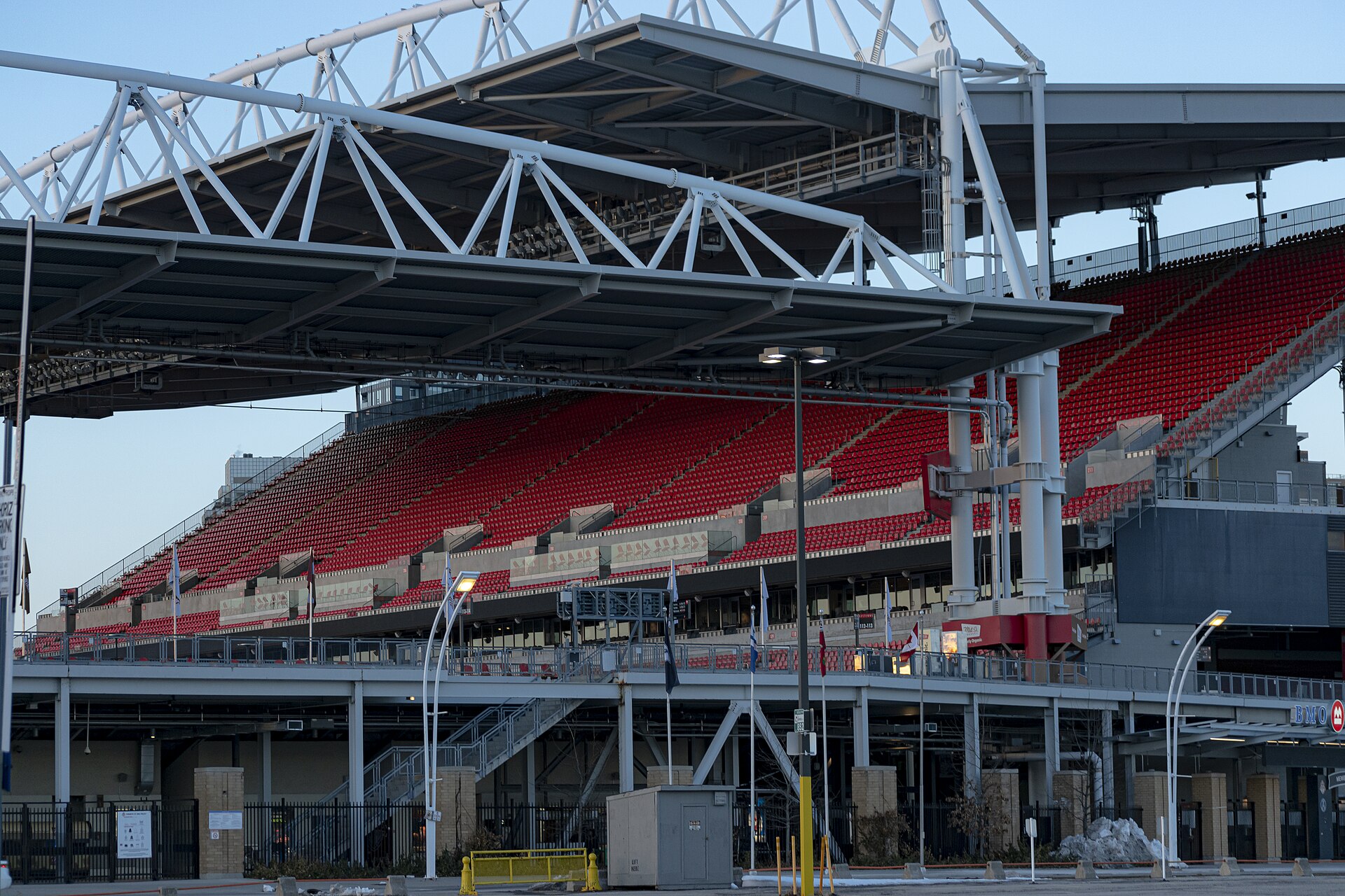 Steep bank of red seats under the white steel canopy of a stadium stand, seen from outside the ground
