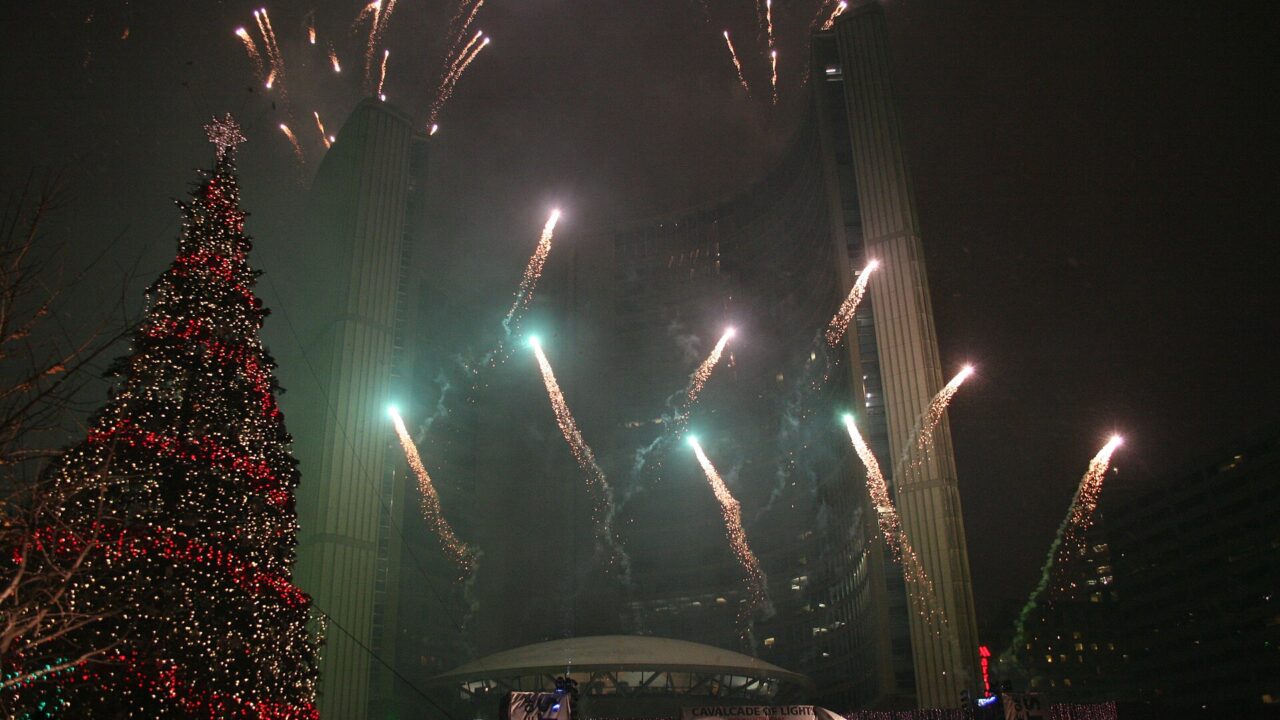 Fireworks bursting over the curved towers of Toronto City Hall at night beside a tall Christmas tree lit in red and white