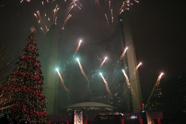 Fireworks bursting over the curved towers of Toronto City Hall at night beside a tall Christmas tree lit in red and white
