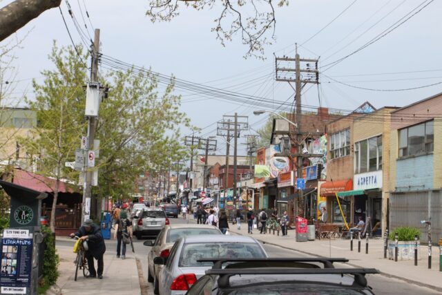 A busy street in Kensington Market on a spring day, with colourful shopfronts, murals, cafe tables on the pavement and people walking under a tangle of overhead wires