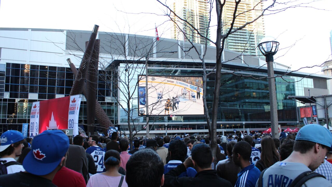 Large crowd in Maple Leafs jerseys watching a hockey game on a giant outdoor screen outside the arena in Toronto