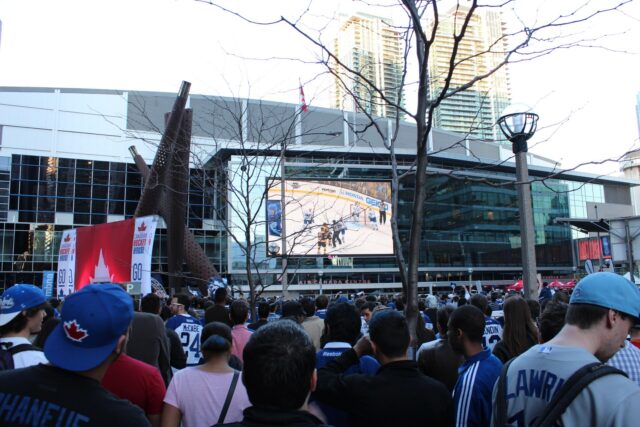 Large crowd in Maple Leafs jerseys watching a hockey game on a giant outdoor screen outside the arena in Toronto