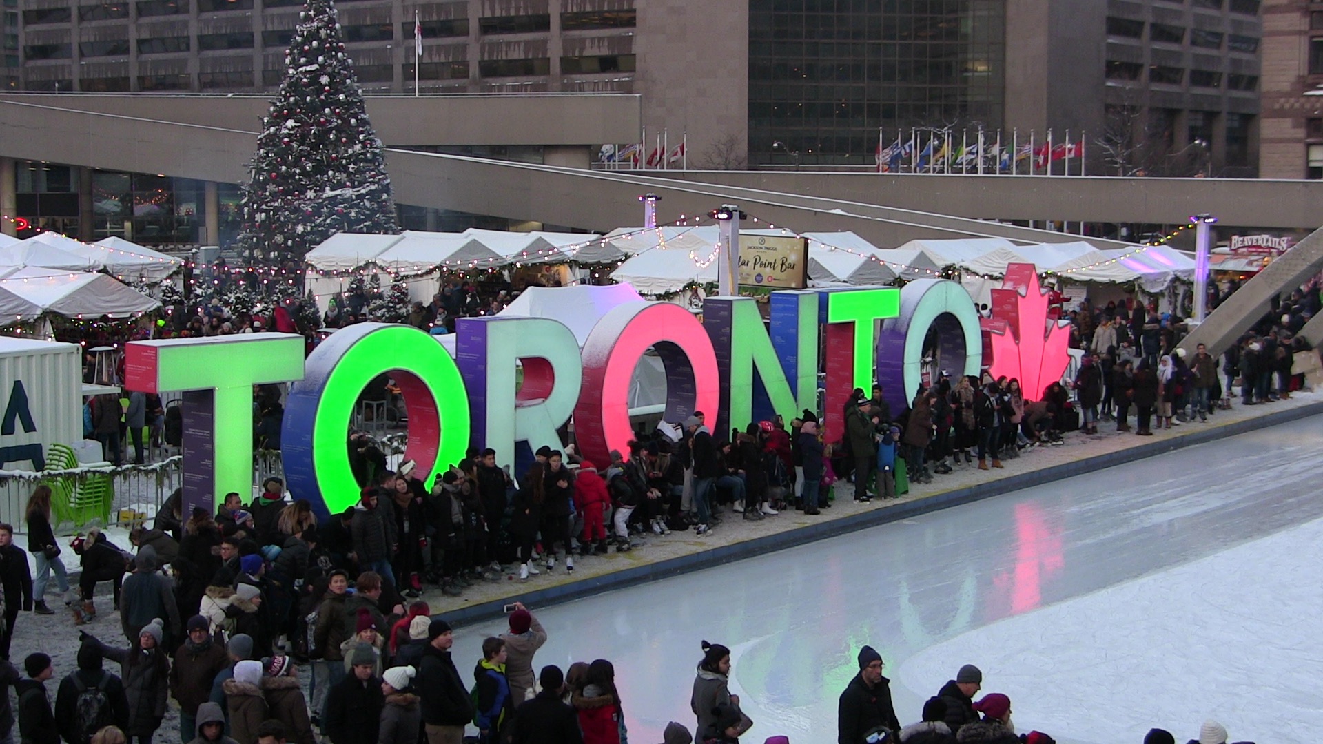 Crowds gathered around the illuminated multicoloured Toronto sign beside the skating rink at Nathan Phillips Square, with a Christmas tree and white market tents behind