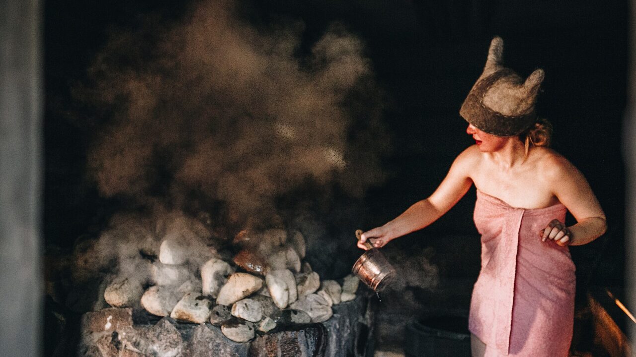 A woman in a towel and felt sauna hat pouring water from a ladle onto hot stones, sending up a cloud of steam in a dark sauna
