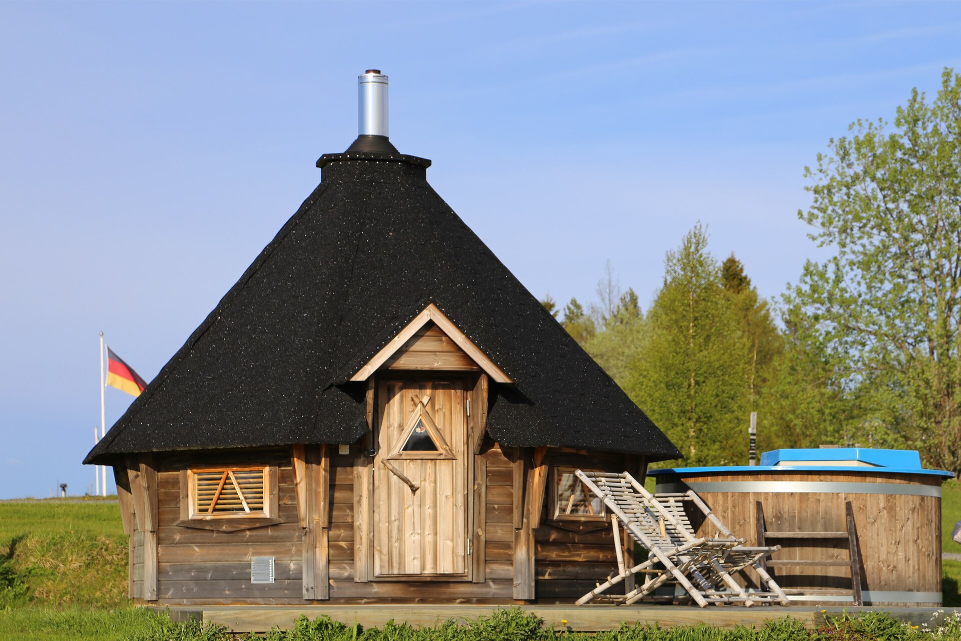 A small wooden hexagonal sauna hut with a black roof and chimney next to a wooden hot tub and lounge chairs on a sunny day