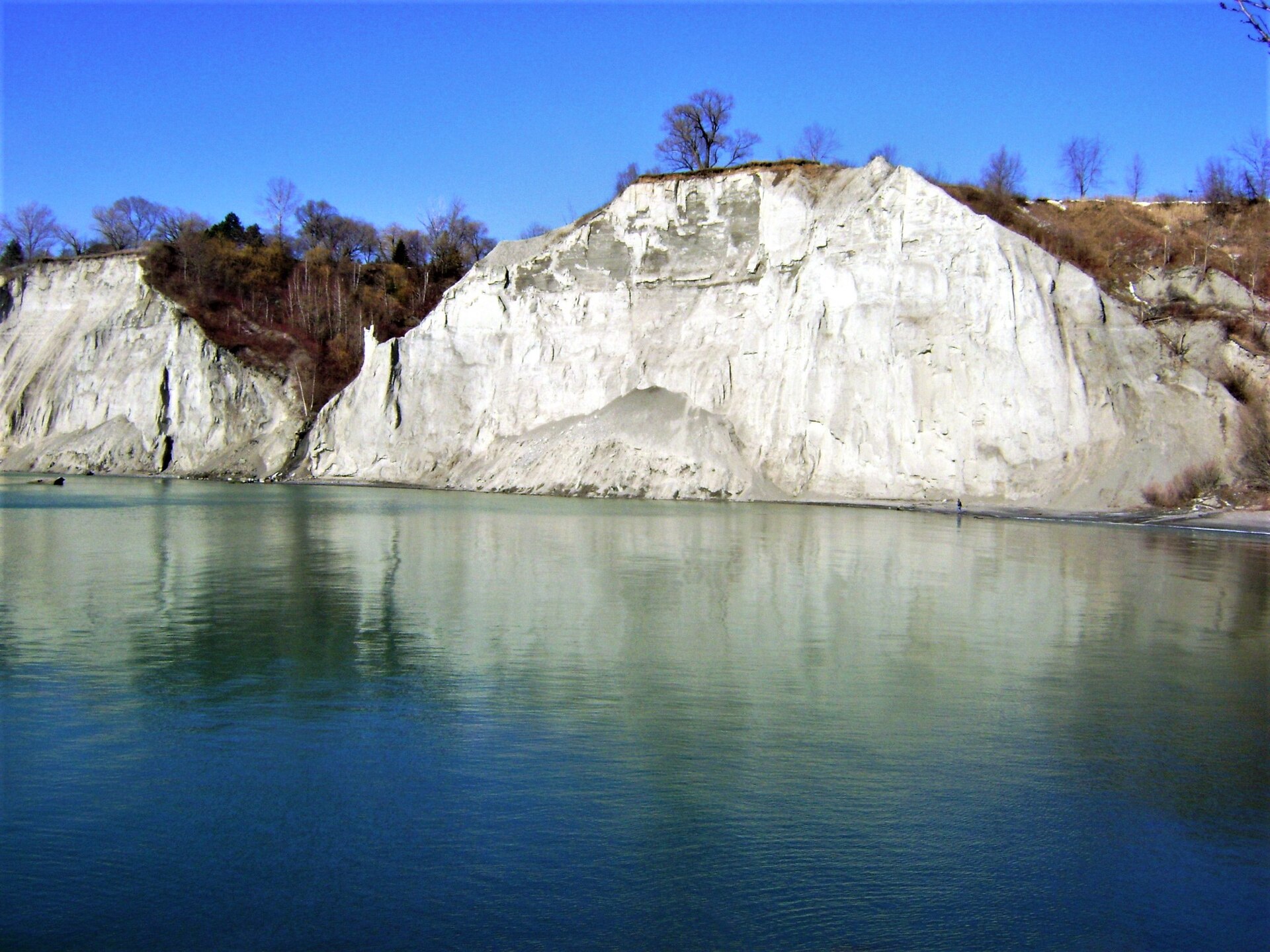 White eroding cliff face rising above the turquoise water of Lake Ontario with bare trees along the top
