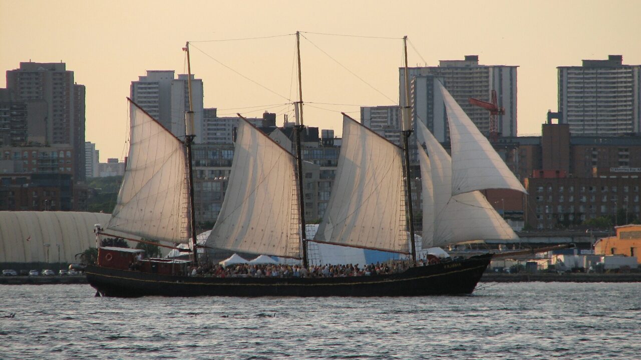 Three-masted tall ship under full sail on Toronto harbour at sunset with the city behind