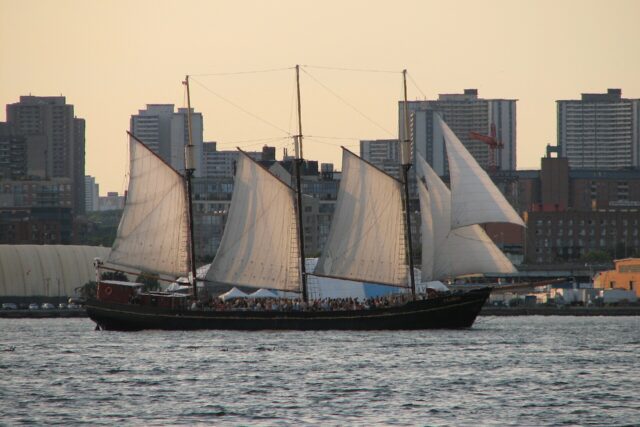 Three-masted tall ship under full sail on Toronto harbour at sunset with the city behind
