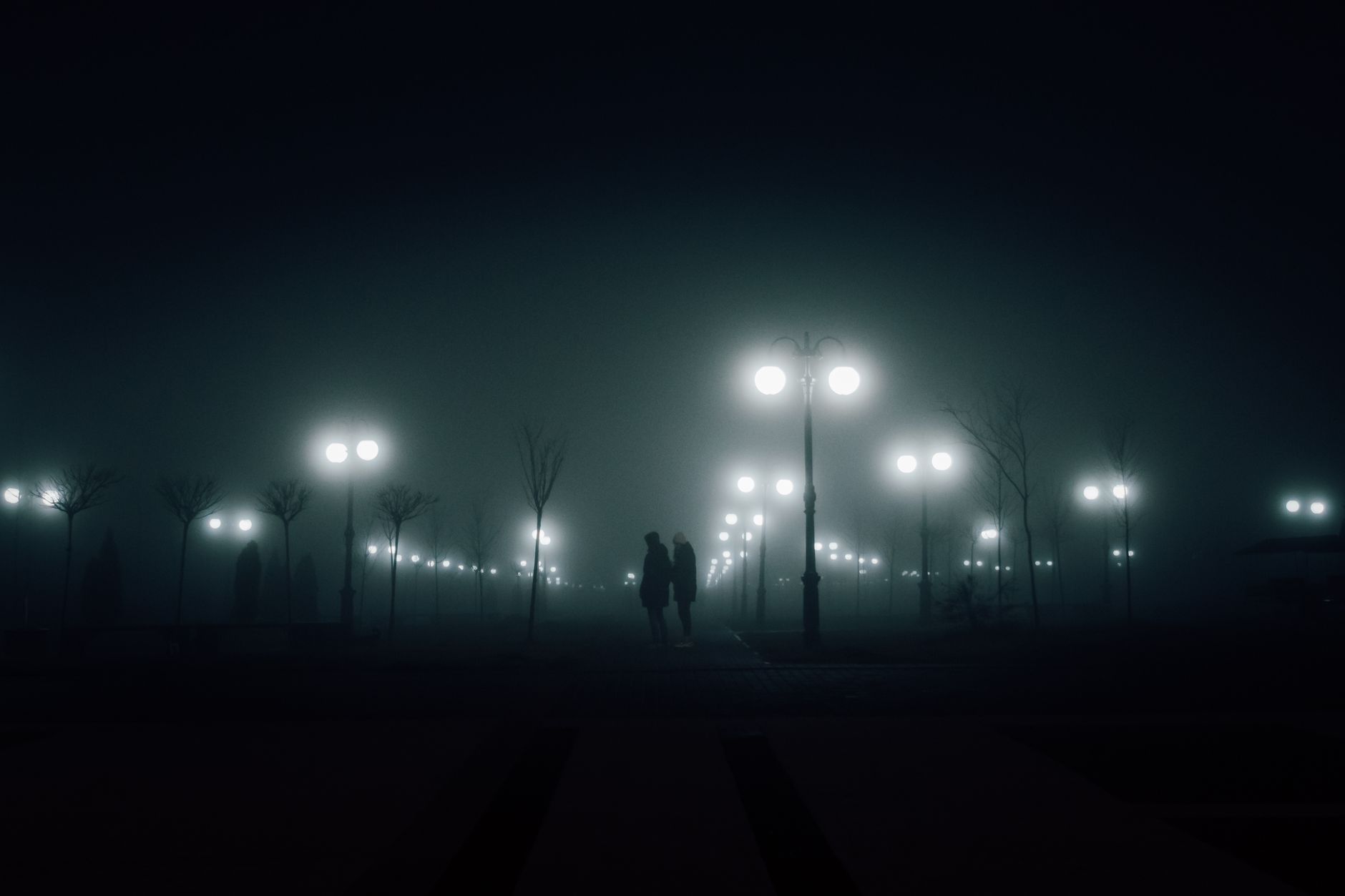 Silhouetted figures walking through a foggy, lamp-lit park after dark