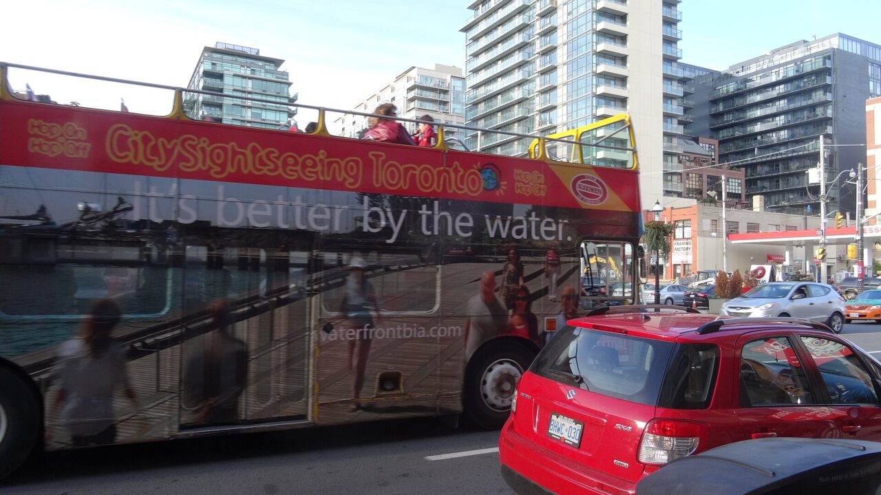 Red open-top double-decker sightseeing bus in downtown Toronto traffic with passengers on the upper deck
