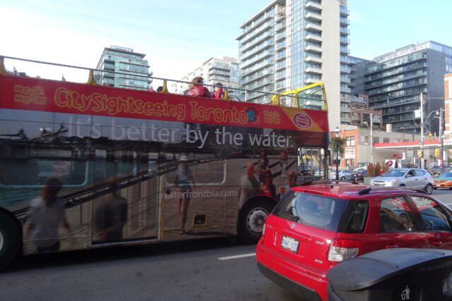 Red open-top double-decker sightseeing bus in downtown Toronto traffic with passengers on the upper deck