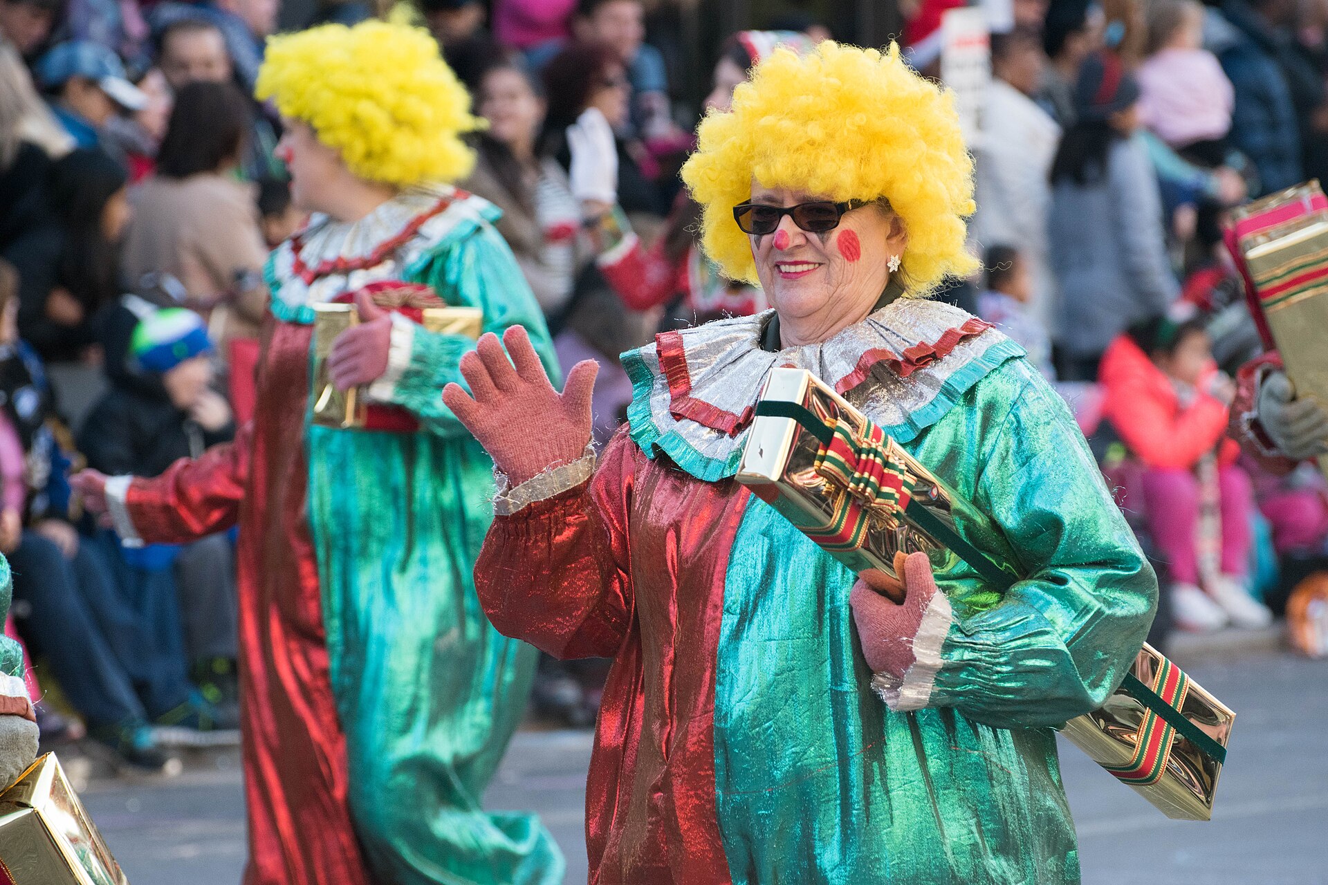 Two parade performers in red and green satin clown costumes and yellow wigs waving to crowds lining a Toronto street