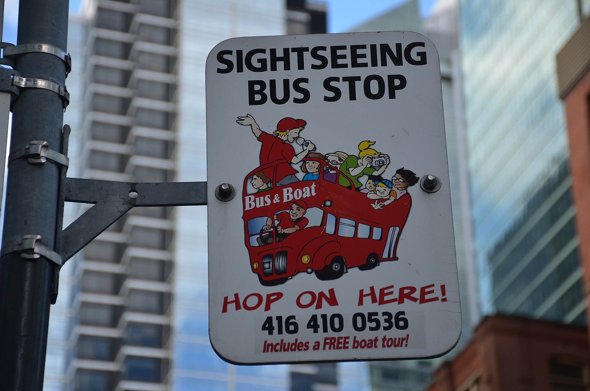 Street sign reading Sightseeing Bus Stop, Hop On Here, mounted on a pole with office towers behind