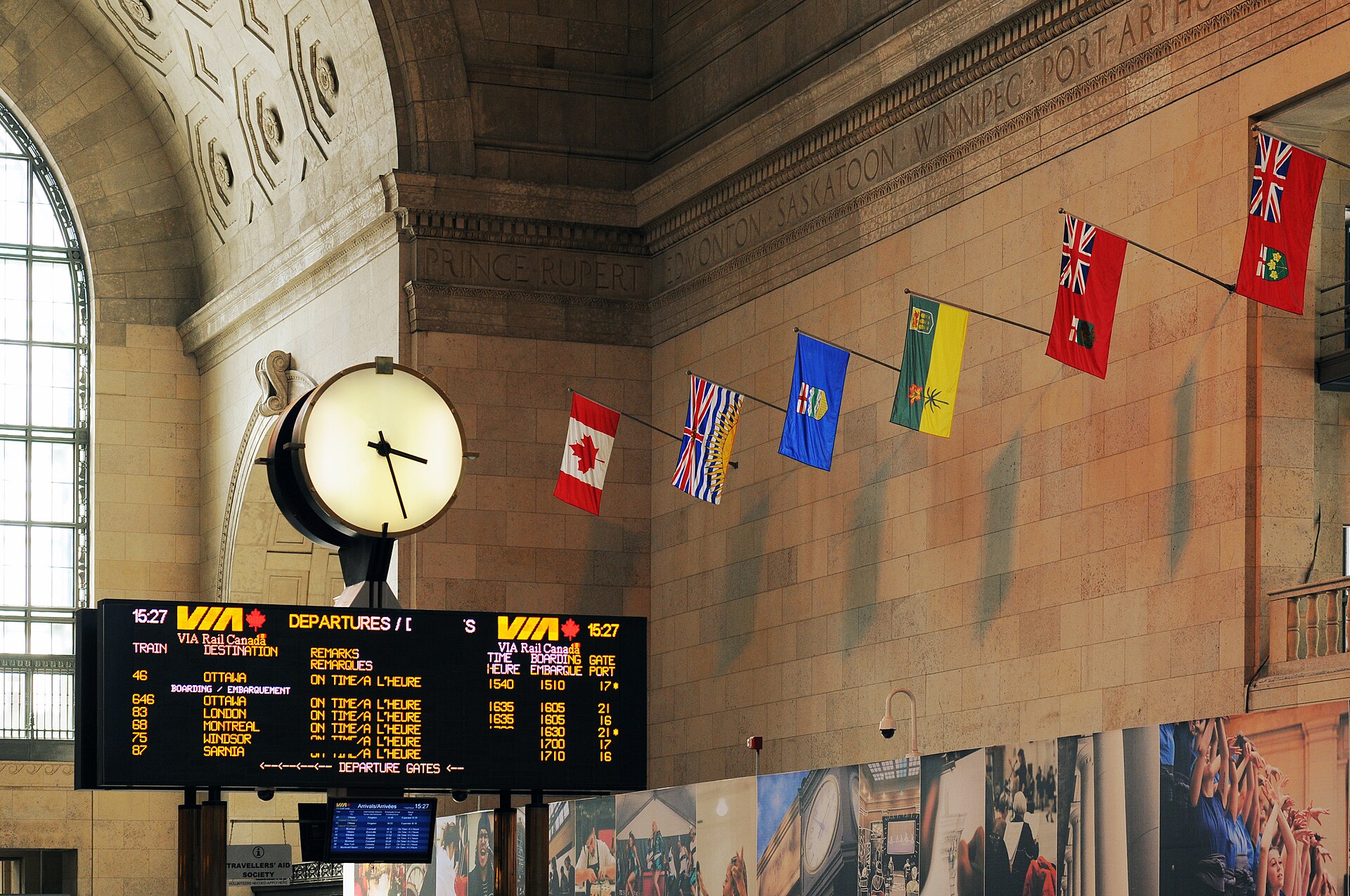 Carved Canadian place names in the stone frieze of Union Station Toronto above a VIA Rail departure board and provincial flags