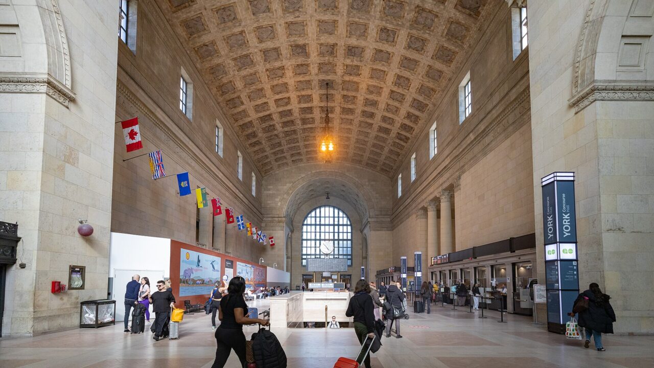The vaulted stone Great Hall of Union Station Toronto with arched windows, hanging provincial flags and travellers with luggage