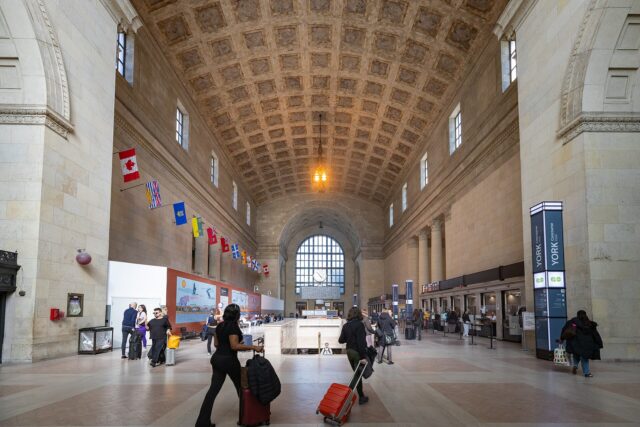 The vaulted stone Great Hall of Union Station Toronto with arched windows, hanging provincial flags and travellers with luggage