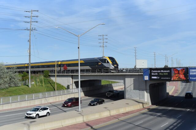A grey and yellow VIA Rail passenger train crossing a concrete road bridge over a multi-lane road on a sunny day