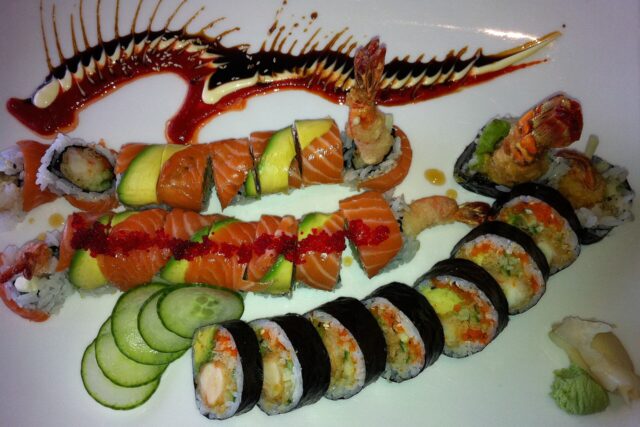 A platter of salmon and avocado topped maki rolls and nori-wrapped shrimp tempura rolls decorated with swirls of eel sauce and mayo