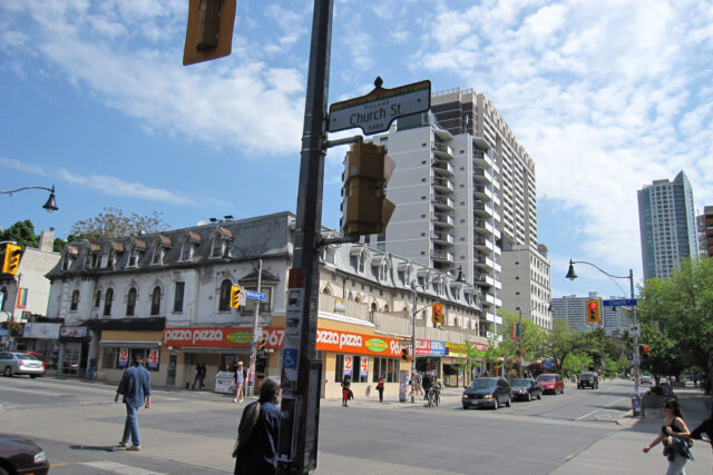 The corner of Church Street and Wellesley Street East in Toronto, with a rainbow-topped Church-Wellesley Village street sign, pedestrians and Victorian buildings on a sunny day
