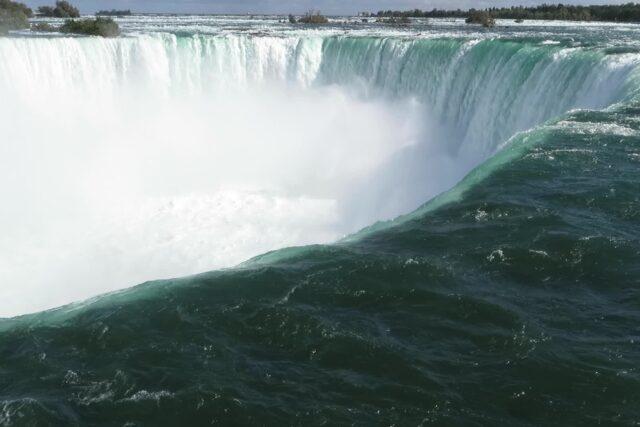Green water curving over the brink of the Horseshoe Falls at Niagara Falls, Ontario, with a cloud of white mist rising below