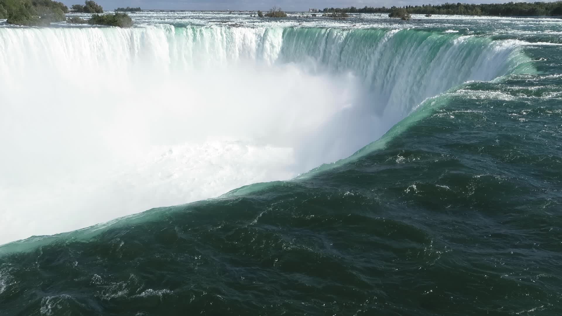 Green water curving over the brink of the Horseshoe Falls at Niagara Falls, Ontario, with a cloud of white mist rising below