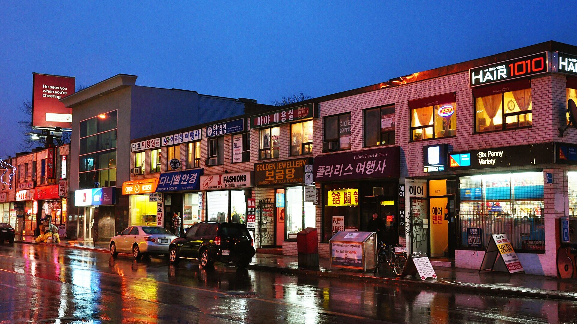 Bloor Street West in Toronto's Koreatown at dusk after rain, with lit Korean-language shop signs and reflections on the wet road