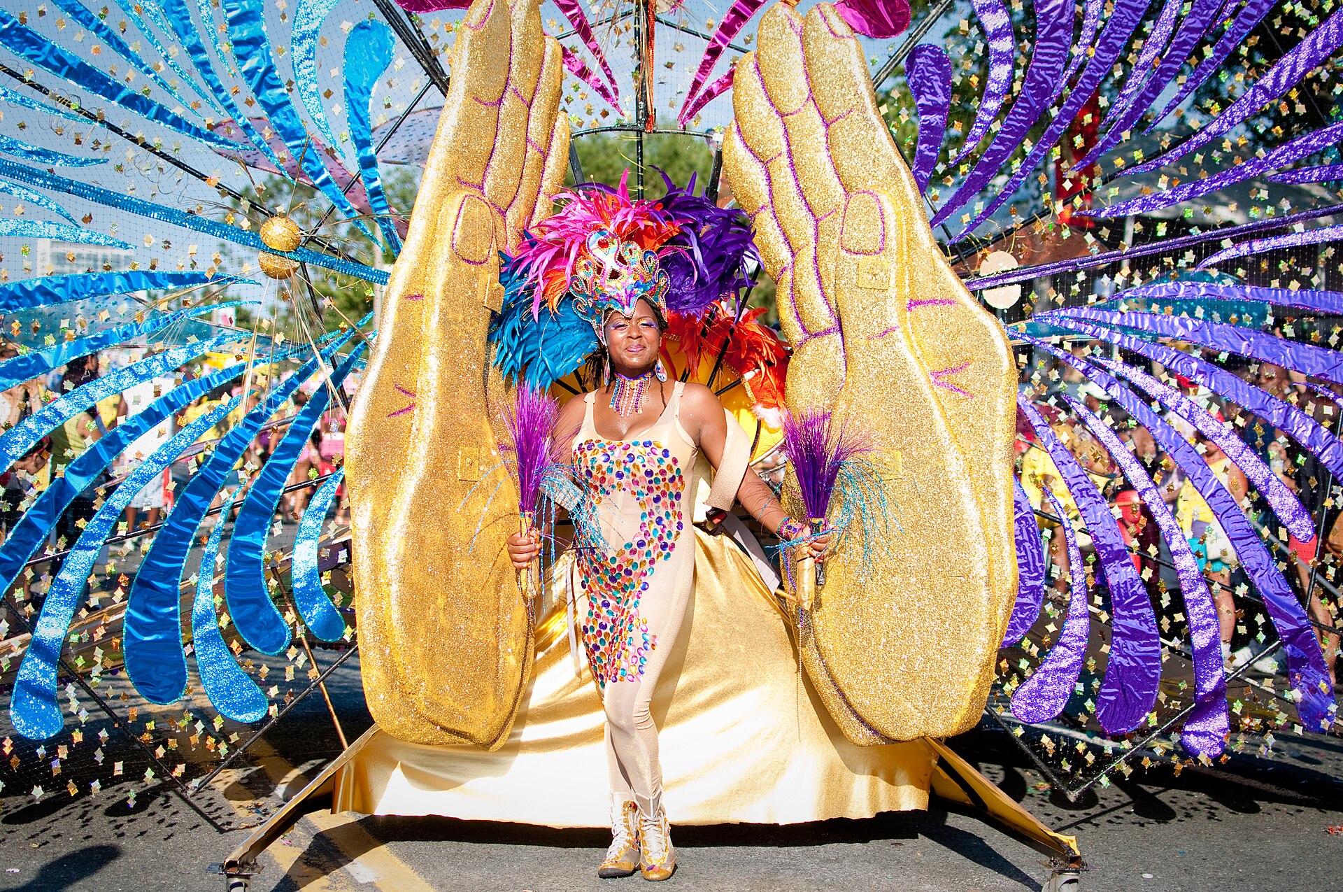 A Toronto Caribana masquerader stands between giant golden hands framed by sweeping blue and purple costume panels.