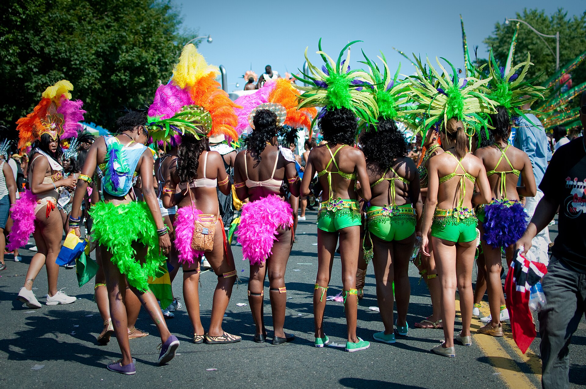 Toronto Caribana participants gather on a sunlit road wearing coordinated green, pink and orange feathered costumes.