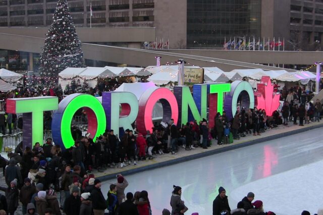 Skaters and visitors crowd the illuminated TORONTO sign beside the rink at Nathan Phillips Square, with a snow-dusted Christmas tree and holiday market tents behind